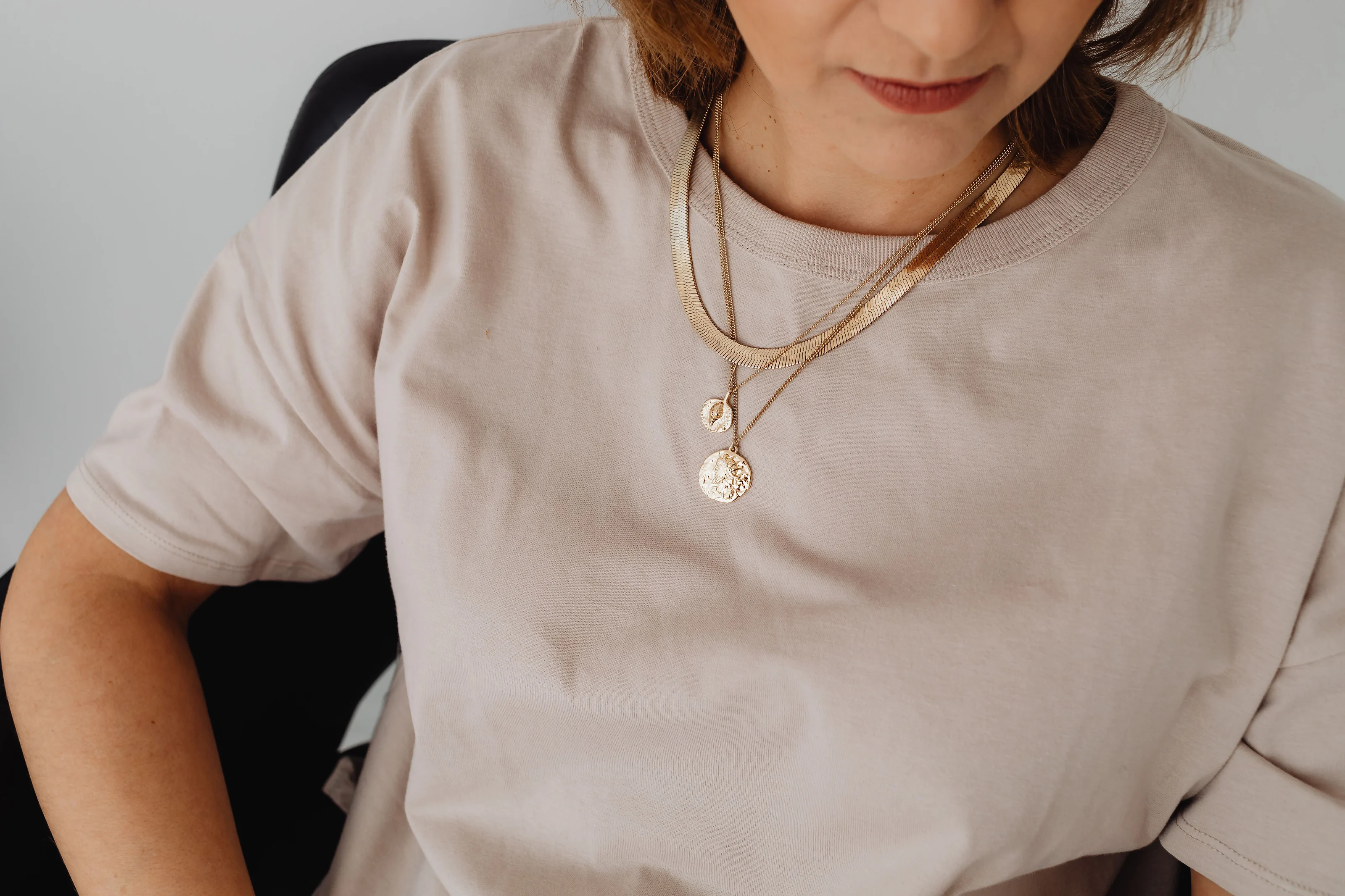 A woman in a soft beige t-shirt wearing layered gold chains and two coin pendants